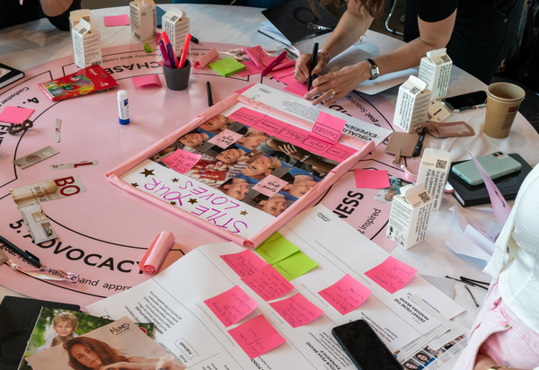 A table is covered with various items, including a pink board game titled ‘THE GAME OF LIFE,’ markers, sticky notes, and printed materials featuring images of people. A person is visible, actively writing or drawing on one of the sticky notes. The scene suggests a creative or planning session involving the board game and other materials